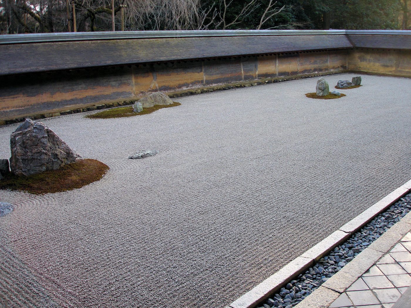 A rectangular garden of finely raked white gravel with a few moss-edged stones set in small groups, enclosed by a low, weathered earthen wall with a tiled top and trees behind.