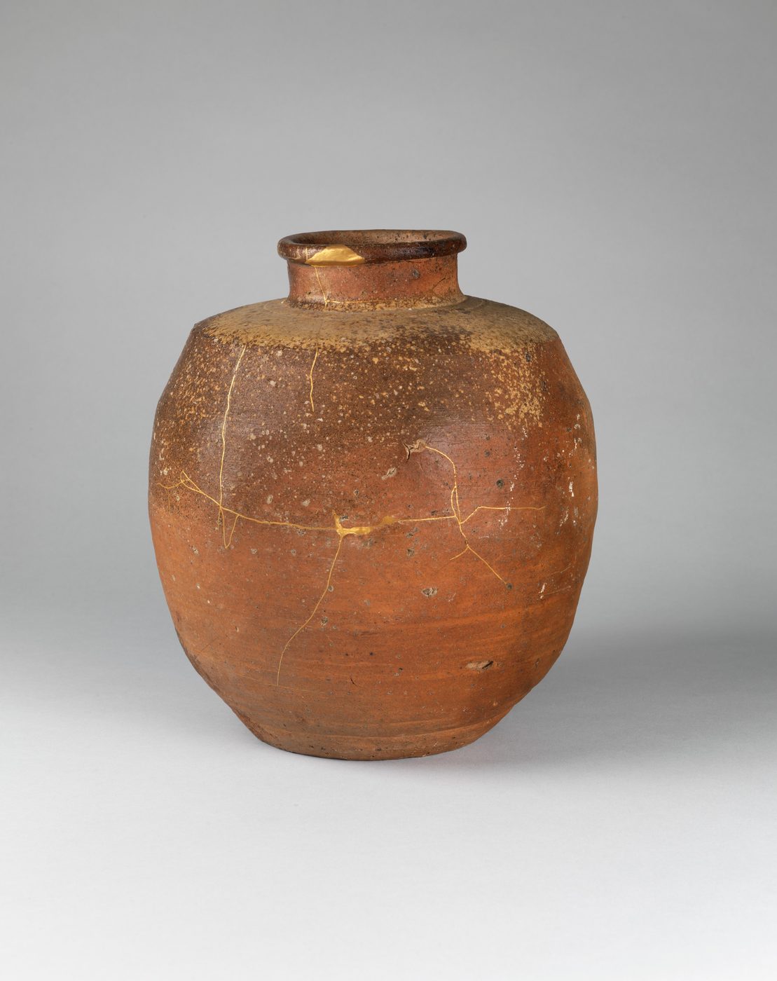 A rounded brown stoneware tea-leaf jar with a short neck, its rough surface crossed by thin, branching seams of gold lacquer where it was broken and repaired.