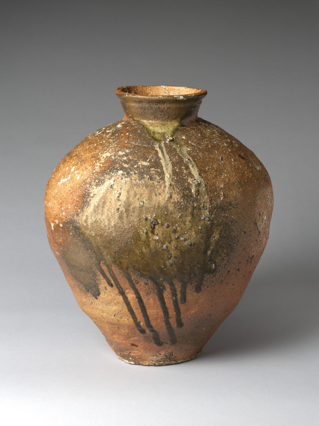 A tall, rounded stoneware storage jar with a rough orange-brown body, a pale crust of melted ash on its shoulder and dark green glaze running down in drips.